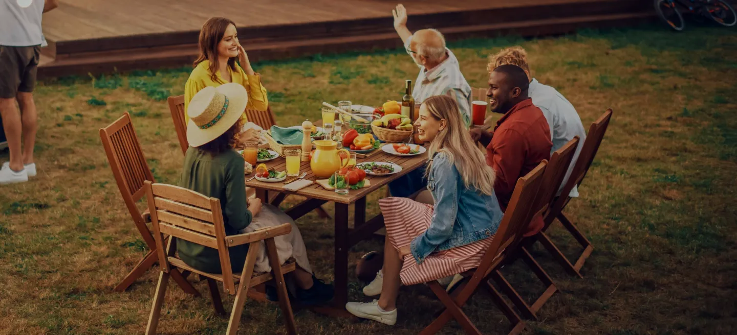 Group of mixed age family members sitting at a table outdoors with food and drinks and smiles on their faces.