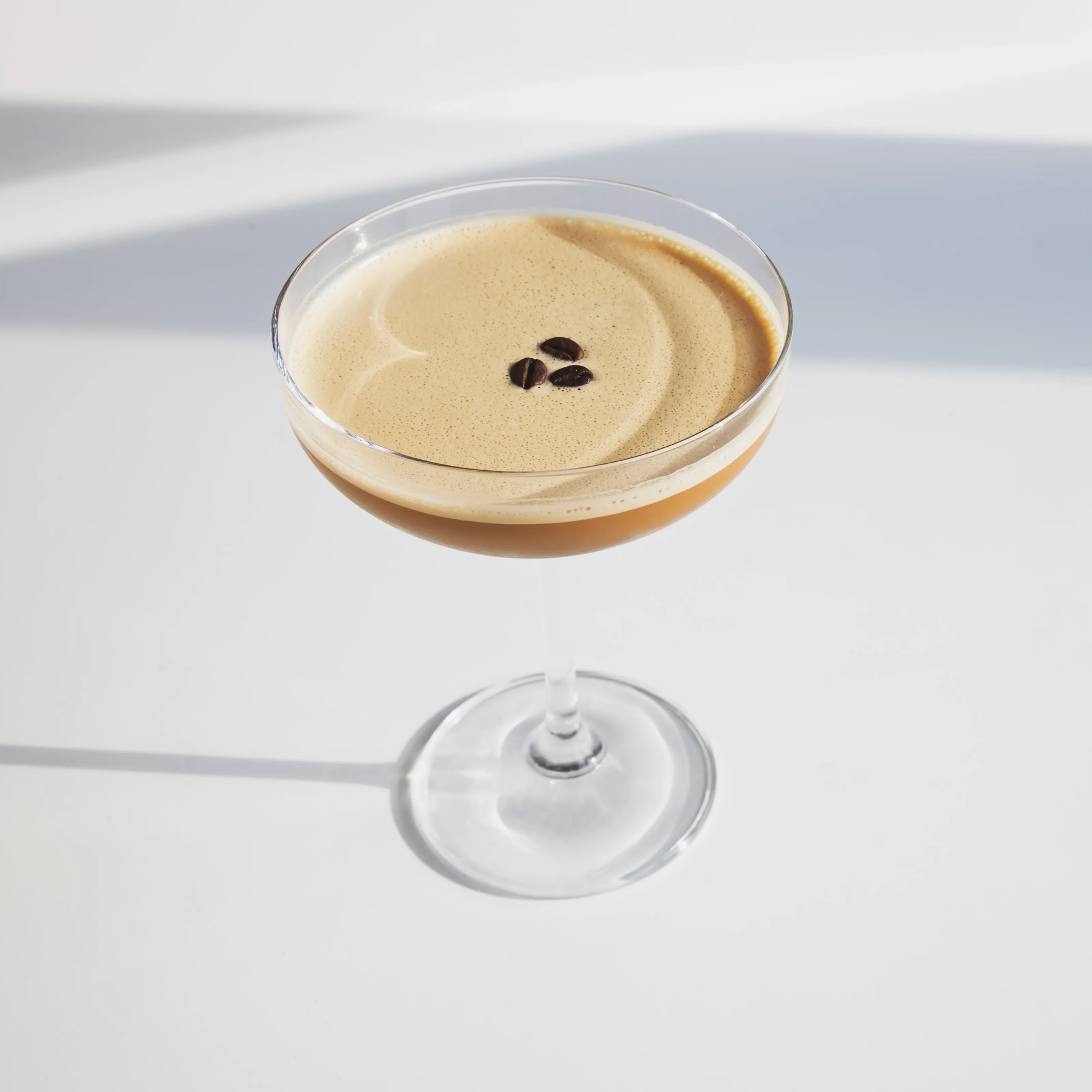 A sleek, coffee colored zero-proof beverage in a coupe glass. A rich foam and 3 coffee beans sit on top.